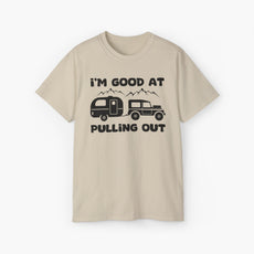 Sand colored t-shirt with humorous text 'I'm good at pulling out' featuring an image of a truck pulling a camping van, set against mountains on a plain background.