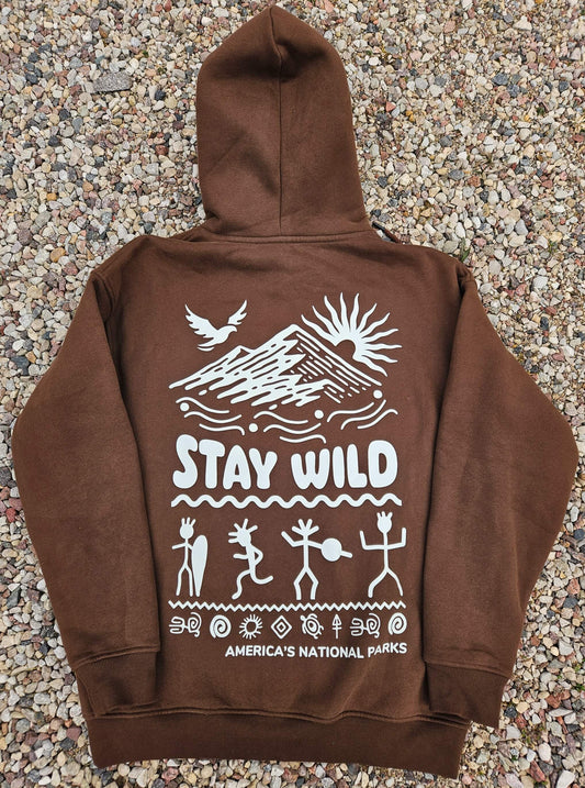 Brown unisex hoodie laid on gravel featuring a white graphic with mountain, sun, bird, petroglyph-style figures, and the phrase "Stay Wild – America's National Parks.