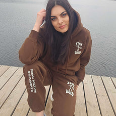 Woman squatting on a wooden dock near a lake, wearing a brown “Stay Wild” hoodie and sweatpants set with white graphics inspired by America’s National Parks.