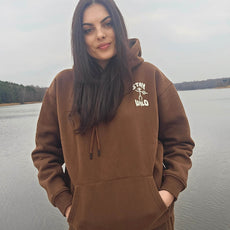 Woman standing outdoors near a lake wearing a matching brown hoodie and sweatpants set with “Stay Wild” and petroglyph-style design inspired by America’s National Parks.