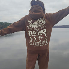 Person standing with arms outstretched facing a calm lake, wearing a brown hoodie with “Stay Wild” and petroglyph-style graphic representing America’s National Parks.