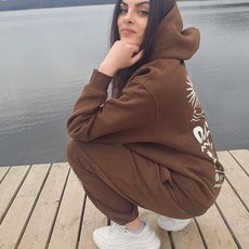 Woman squatting on a wooden dock near a lake, wearing a brown “Stay Wild” hoodie and sweatpants set with white graphics inspired by America’s National Parks.