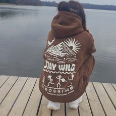 Woman squatting on a wooden dock near a lake, wearing a brown “Stay Wild” hoodie and sweatpants set with white graphics inspired by America’s National Parks.