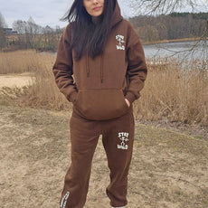 Woman wearing a brown “Stay Wild” hoodie and sweatpants set with white petroglyph graphics, standing outdoors by a lake and dry grass on a cloudy day.