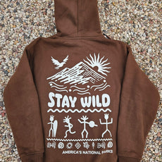 Brown unisex hoodie laid on gravel featuring a white graphic with mountain, sun, bird, petroglyph-style figures, and the phrase 