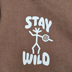 Close-up of a brown hoodie featuring a white DTF print of a stick-figure petroglyph holding a spiral design with the words “STAY WILD” above and below.