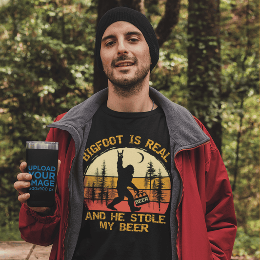 Man wearing a black t-shirt with the text 'Bigfoot is real and he stole my beer,' featuring a Bigfoot holding a beer, standing in a nature camping setting.