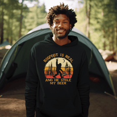 Man wearing a black hoodie with the text 'Bigfoot is real and he stole my beer,' featuring a Bigfoot holding a beer, standing in a nature camping setting